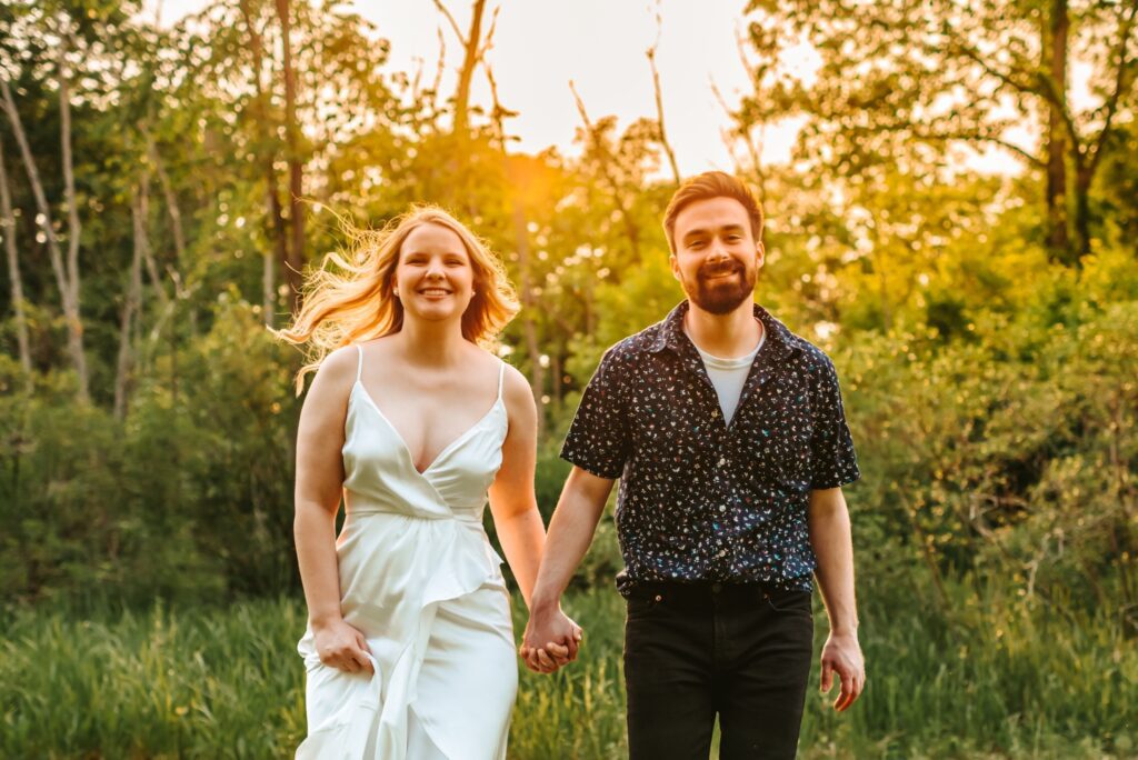 engaged couple running together in a field with trees