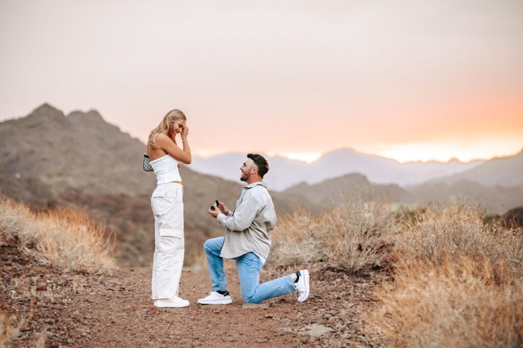 man on one knee, proposing to girlfriend at sunset with mountains in the background
